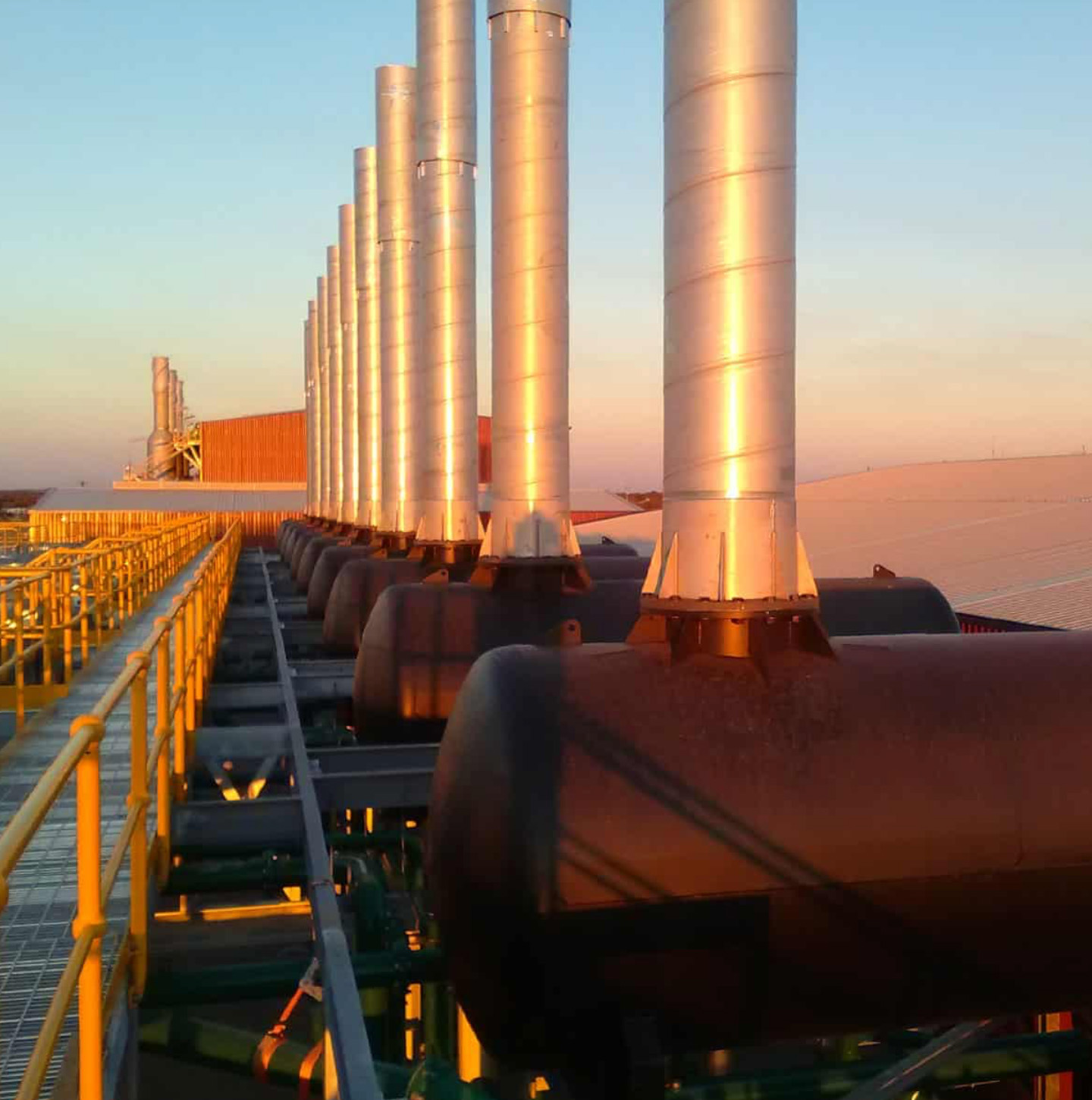 Row of stainless-steel exhaust stacks and silencers on a power generation plant at sunset